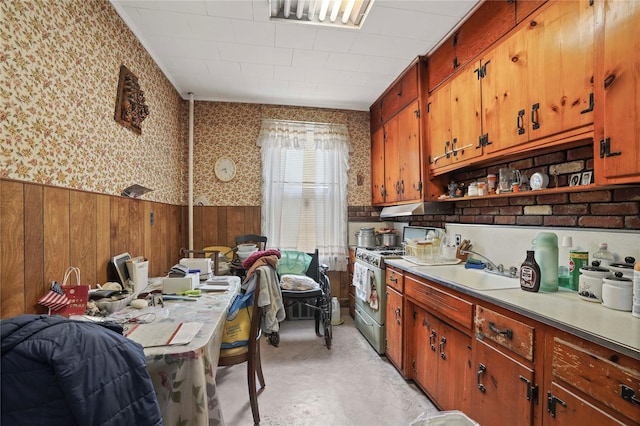 kitchen with brown cabinetry, wainscoting, stainless steel gas stove, wallpapered walls, and light countertops