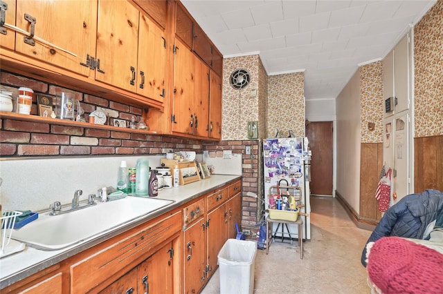 kitchen featuring brown cabinetry, wainscoting, wallpapered walls, light countertops, and a sink