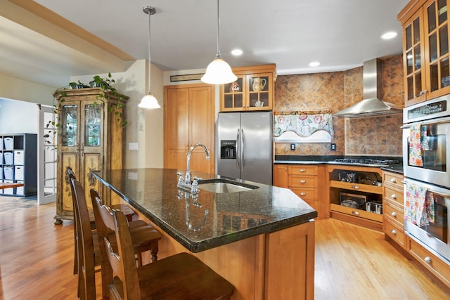 kitchen with wall chimney range hood, a sink, light wood-style flooring, brown cabinetry, and appliances with stainless steel finishes