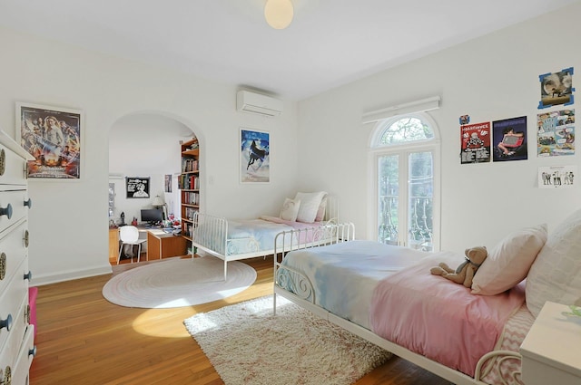 bedroom with arched walkways, baseboards, an AC wall unit, and wood finished floors