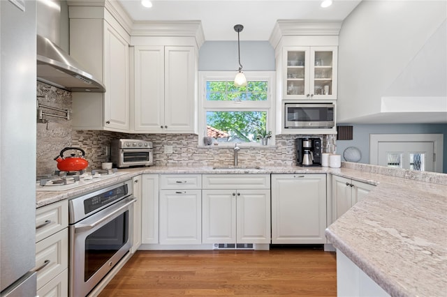 kitchen featuring wall chimney exhaust hood, white cabinetry, a sink, light wood-style flooring, and appliances with stainless steel finishes