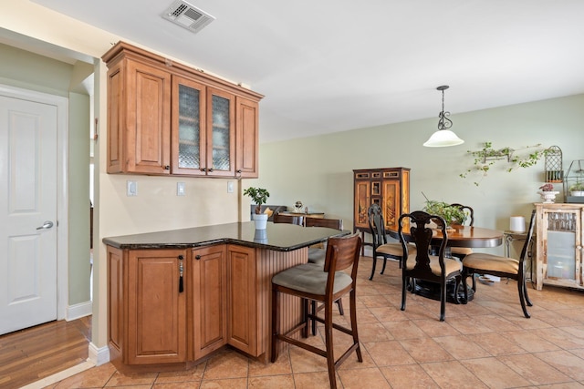 kitchen featuring visible vents, glass insert cabinets, brown cabinets, a breakfast bar, and hanging light fixtures