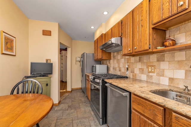 kitchen featuring stone tile floors, under cabinet range hood, stainless steel appliances, tasteful backsplash, and brown cabinetry