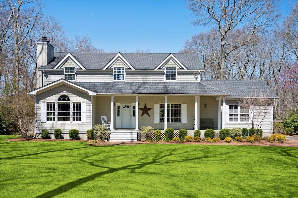 view of front facade with a front yard, roof with shingles, a chimney, and covered porch