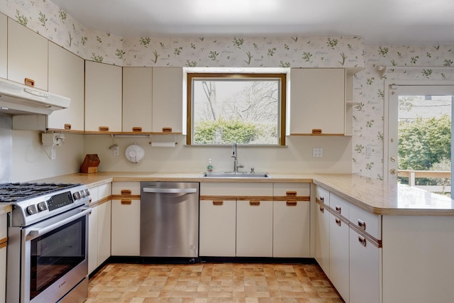 kitchen with a sink, appliances with stainless steel finishes, light countertops, wallpapered walls, and a peninsula