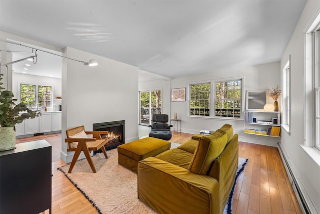 living room with wood-type flooring, a warm lit fireplace, a baseboard heating unit, and a healthy amount of sunlight