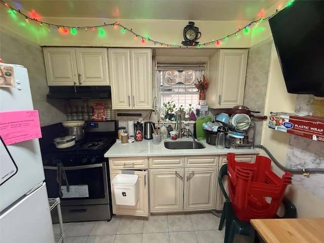 kitchen with stainless steel gas range oven, under cabinet range hood, a sink, light tile patterned floors, and freestanding refrigerator
