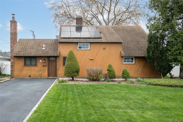 view of front of home with a chimney, a shingled roof, roof mounted solar panels, a front yard, and aphalt driveway