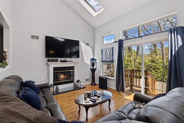 living area with high vaulted ceiling, a glass covered fireplace, wood finished floors, a skylight, and visible vents