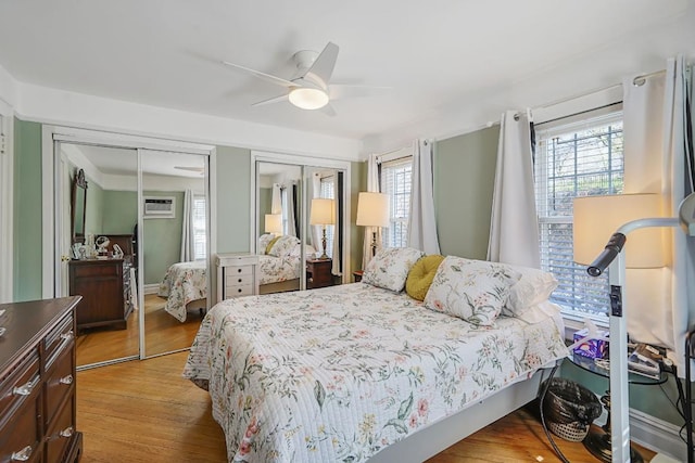 bedroom with a ceiling fan, a wall unit AC, multiple closets, and light wood-style floors