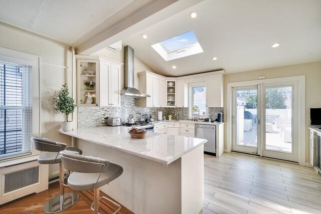 kitchen featuring wall chimney exhaust hood, a peninsula, white cabinets, backsplash, and appliances with stainless steel finishes