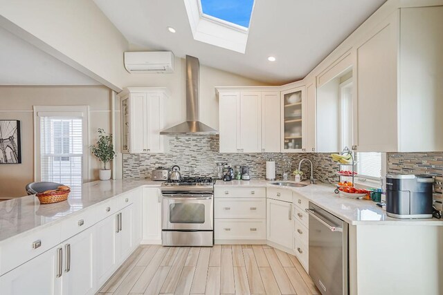 kitchen with wall chimney range hood, vaulted ceiling with skylight, a sink, a wall mounted AC, and stainless steel appliances