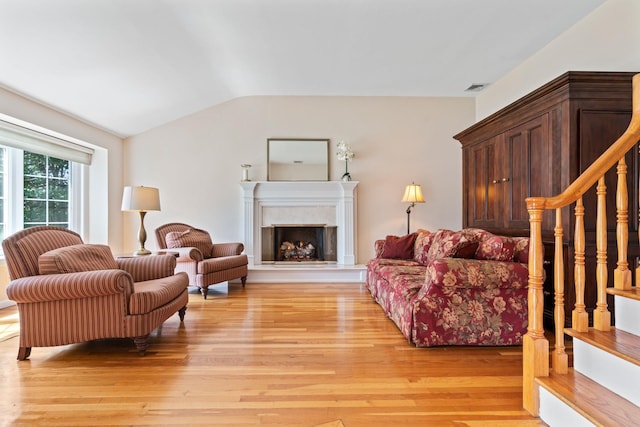 living area with light wood finished floors, vaulted ceiling, visible vents, a fireplace with raised hearth, and stairs