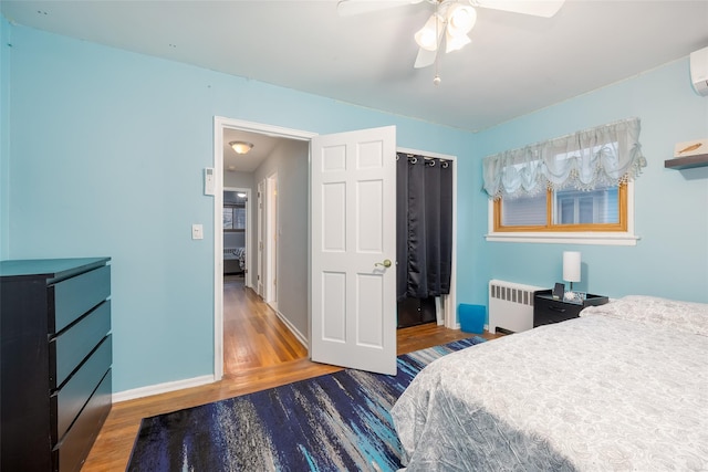 bedroom with radiator heating unit, wood finished floors, a ceiling fan, an AC wall unit, and baseboards
