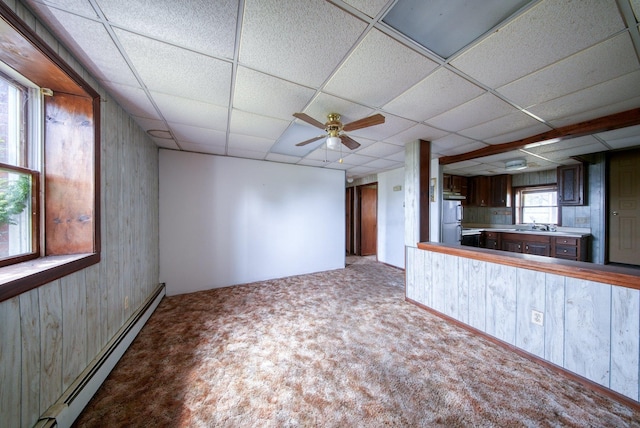 unfurnished living room featuring ceiling fan, wooden walls, a paneled ceiling, a baseboard heating unit, and carpet