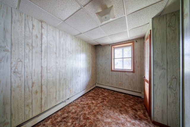 empty room with a baseboard radiator, dark colored carpet, a drop ceiling, and wood walls