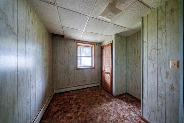 carpeted empty room with wooden walls, baseboards, a baseboard heating unit, and a drop ceiling