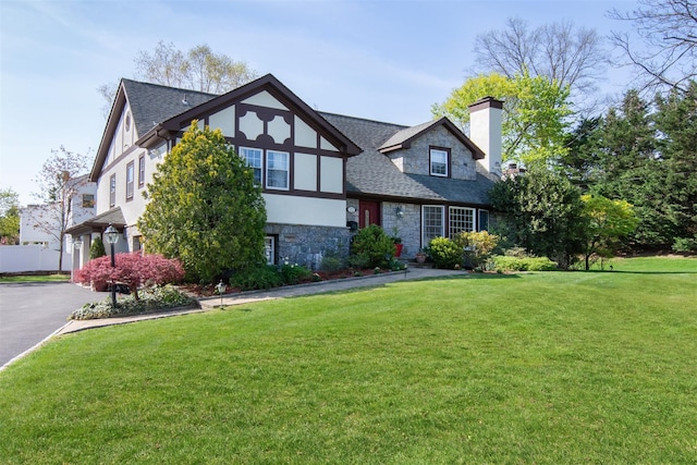 tudor-style house with a front yard, a shingled roof, stone siding, a chimney, and stucco siding