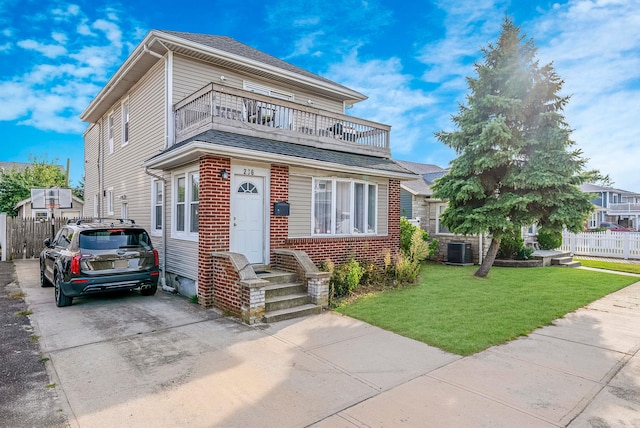 view of front facade featuring central air condition unit, fence, brick siding, a balcony, and a front yard