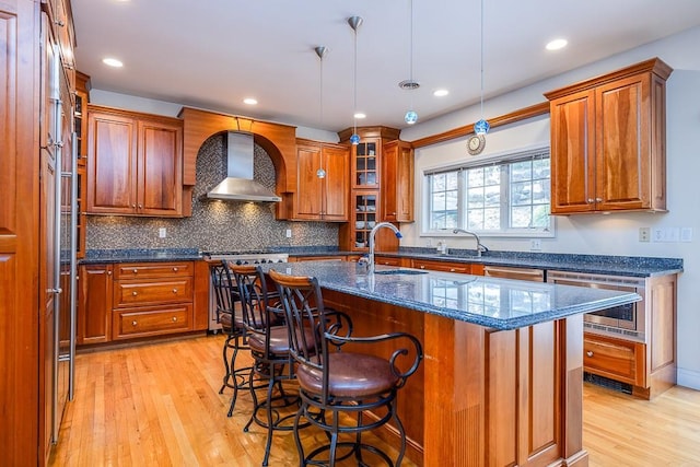 kitchen featuring light wood-style floors, wall chimney range hood, brown cabinets, a sink, and range