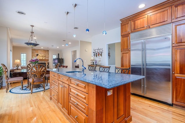 kitchen with a sink, light wood-style floors, brown cabinetry, dark stone countertops, and stainless steel built in fridge