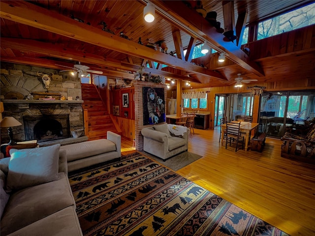 living room with a ceiling fan, wood finished floors, a stone fireplace, wood walls, and beam ceiling