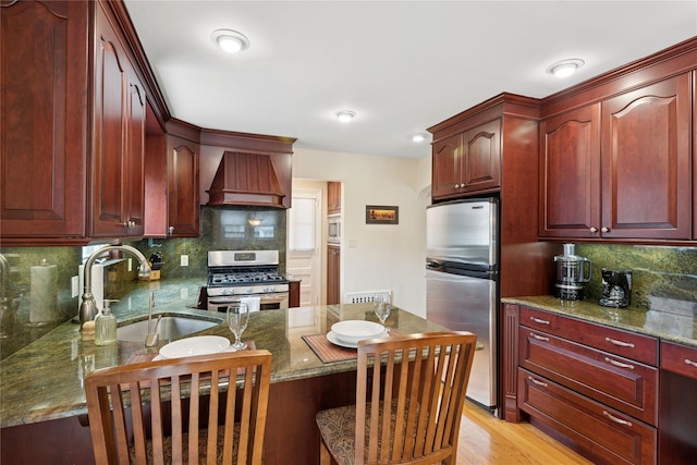 kitchen with reddish brown cabinets, a sink, premium range hood, decorative backsplash, and appliances with stainless steel finishes