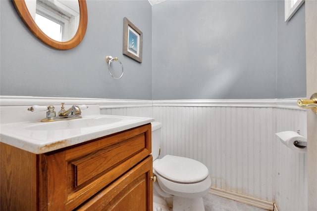 half bathroom with wainscoting, vanity, tile patterned flooring, and toilet