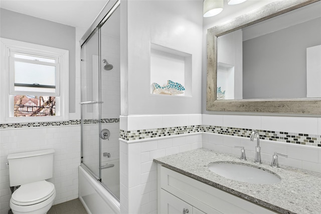 bathroom with a wainscoted wall, enclosed tub / shower combo, toilet, vanity, and tile walls