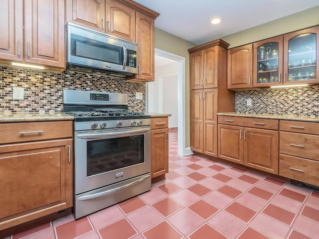 kitchen featuring glass insert cabinets, brown cabinets, stainless steel appliances, backsplash, and light stone counters