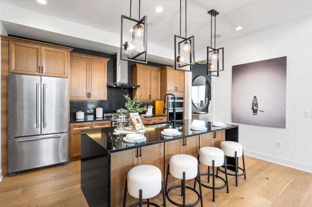 kitchen with an island with sink, light wood-style flooring, wall chimney range hood, dark countertops, and high quality fridge