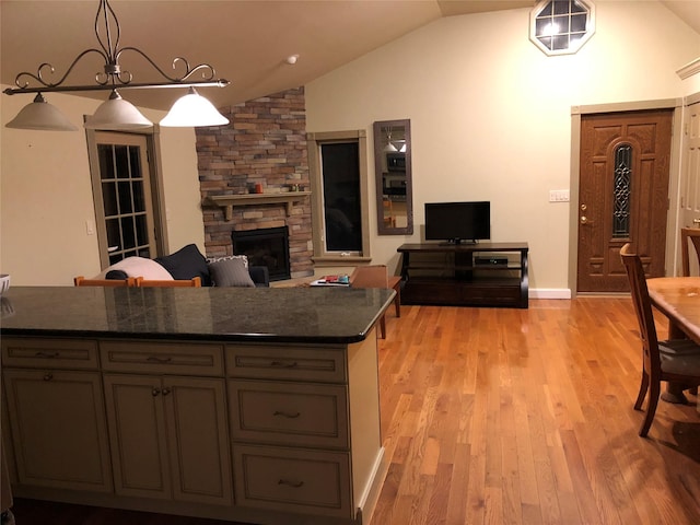 kitchen with light wood-style flooring, decorative light fixtures, vaulted ceiling, dark countertops, and a stone fireplace