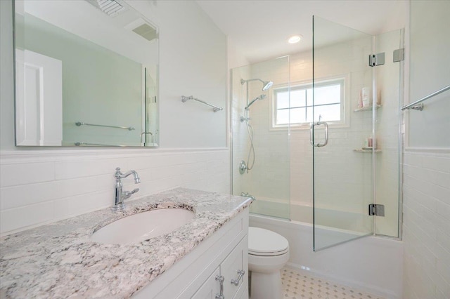 bathroom with tile walls, vanity, toilet, a wainscoted wall, and combined bath / shower with glass door
