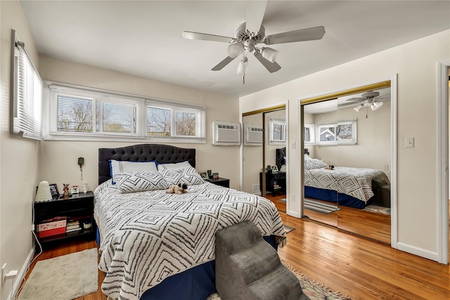 bedroom featuring a wall unit AC, a ceiling fan, wood finished floors, baseboards, and two closets