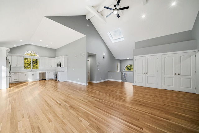 unfurnished living room featuring light wood-type flooring, beam ceiling, a skylight, high vaulted ceiling, and ceiling fan