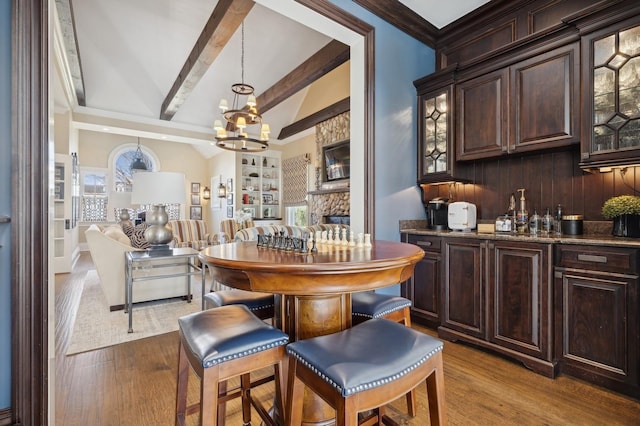 bar featuring a chandelier, light wood-style floors, a stone fireplace, and vaulted ceiling with beams