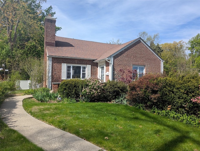 view of front facade with brick siding, roof with shingles, a front yard, and a chimney