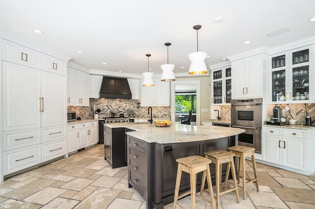 kitchen featuring stainless steel double oven, a kitchen island with sink, custom exhaust hood, stone tile floors, and decorative backsplash