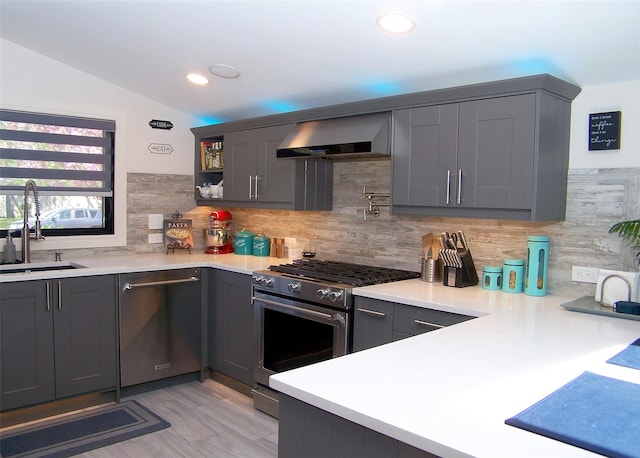 kitchen featuring wall chimney range hood, backsplash, appliances with stainless steel finishes, a sink, and gray cabinetry