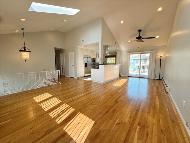 unfurnished living room with ceiling fan, a baseboard heating unit, light wood-type flooring, high vaulted ceiling, and a skylight