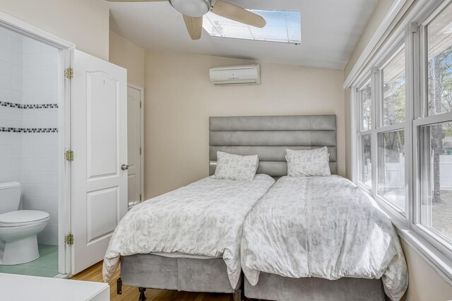 bedroom with ceiling fan, lofted ceiling with skylight, an AC wall unit, wood finished floors, and ensuite bathroom