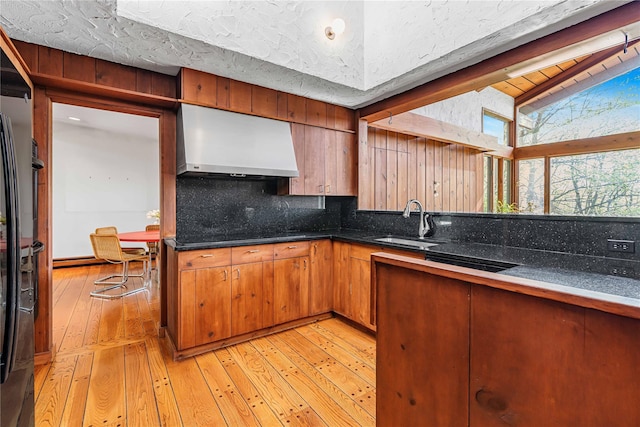 kitchen with wall chimney range hood, a sink, tasteful backsplash, light wood finished floors, and brown cabinetry