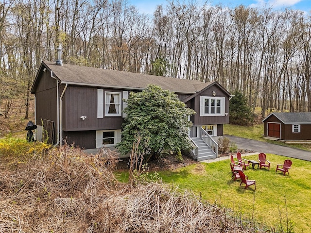 split foyer home featuring a front lawn, stairway, an outdoor structure, driveway, and roof with shingles