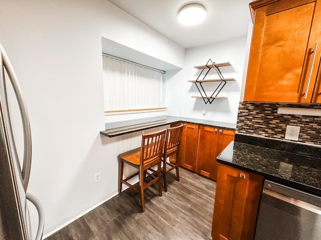 kitchen featuring brown cabinetry, backsplash, wood finished floors, dark stone countertops, and stainless steel appliances