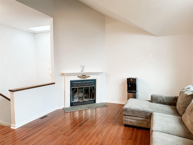 living room featuring a glass covered fireplace, baseboards, wood finished floors, visible vents, and lofted ceiling with skylight