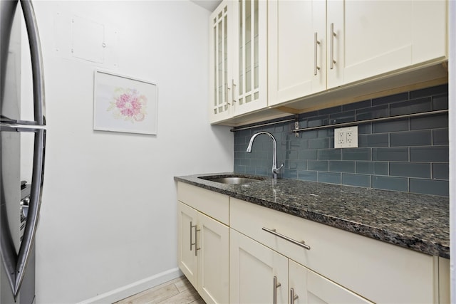 kitchen with glass insert cabinets, dark stone counters, decorative backsplash, a sink, and baseboards