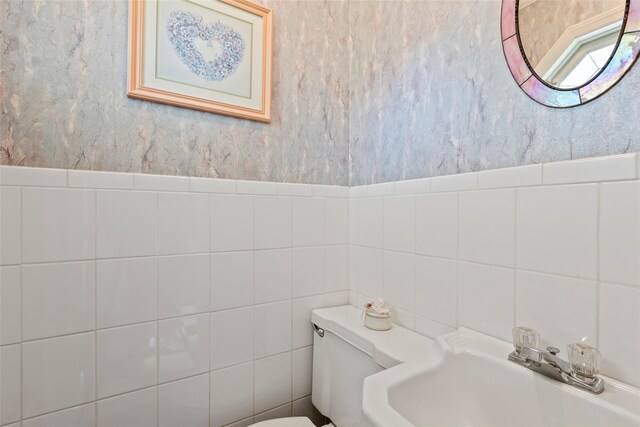 bathroom with toilet, wallpapered walls, tile walls, and a sink