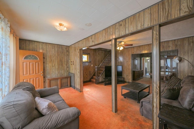 living area featuring carpet floors, stairs, ceiling fan, and wooden walls