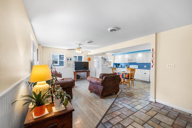 living room featuring baseboards, a ceiling fan, wainscoting, and stone tile floors