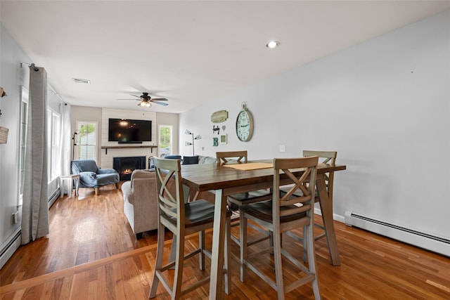 dining room featuring visible vents, a large fireplace, a ceiling fan, wood finished floors, and a baseboard heating unit